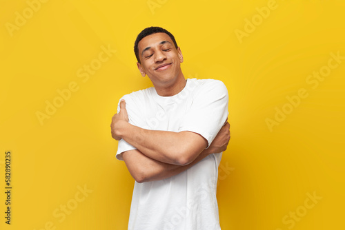 young narcissistic african american guy in white t-shirt hugs himself on yellow isolated background, lonely man photo