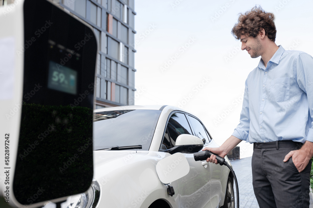 Progressive businessman insert charger plug from charging station to his electric vehicle with apartment condo building in background. Eco friendly rechargeable car powered by sustainable energy. - obrazy, fototapety, plakaty 