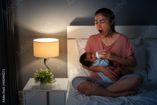 Obraz w ramie sleepy mother yawning and feeding milk bottle to newborn baby on bed at night