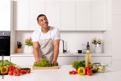 young pensive african american man in apron prepares vegetarian salad with vegetables and greens