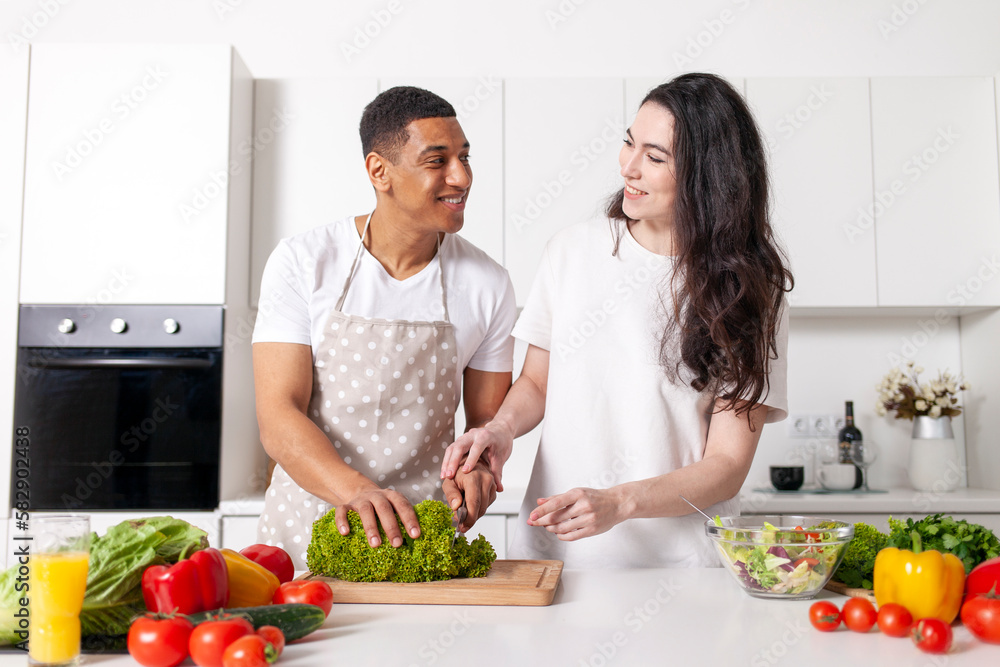 young multiracial couple prepares veggie salad in white modern kitchen, african american guy in apron cuts greens