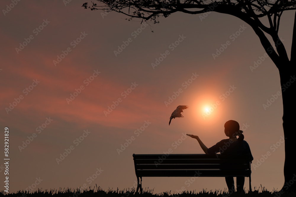 Silhouette of a young woman sitting in a garden chair under a big tree with birds flying towards outstretched hands in the morning.