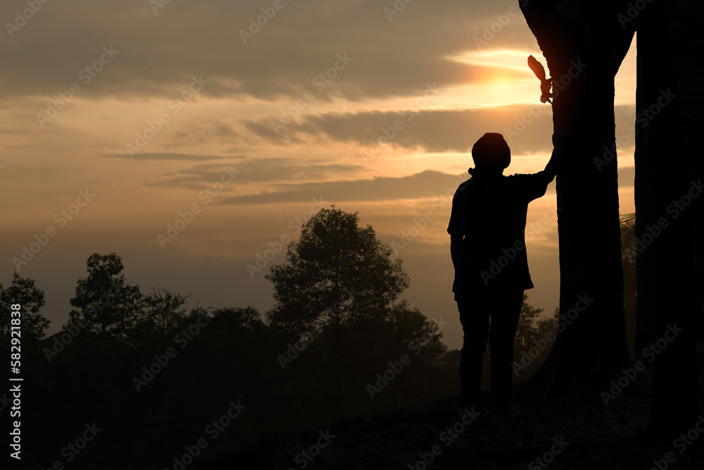 A silhouette of a young woman standing outstretched by a hand at a big tree with a squirrel on the tree in the morning light.