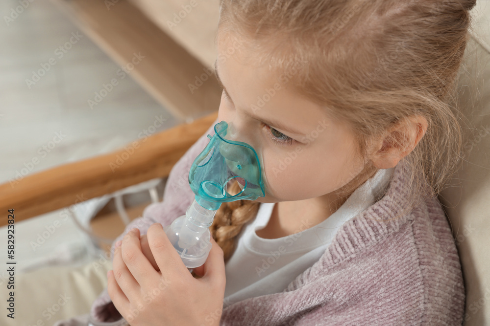 Little girl using nebulizer for inhalation in armchair at home