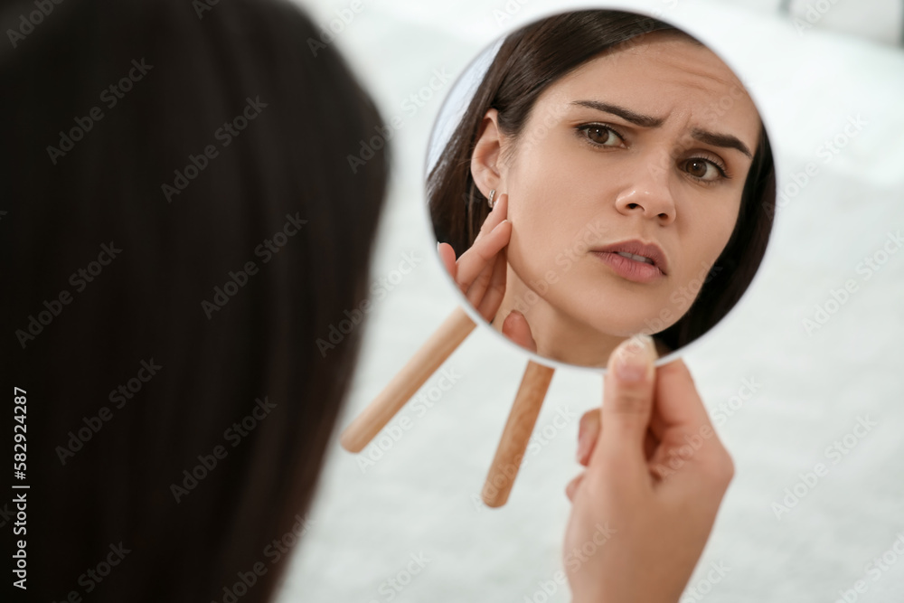 Young woman looking in mirror and touching her face indoors. Hormonal ...