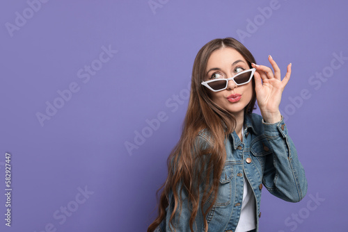 Beautiful young woman with sunglasses blowing kiss on purple background, space for text
