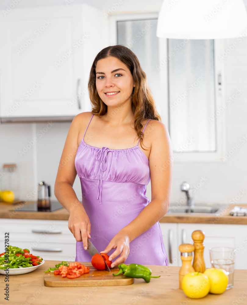 Smiling young woman in nightie cooking vegetable salad in kitchen at home.