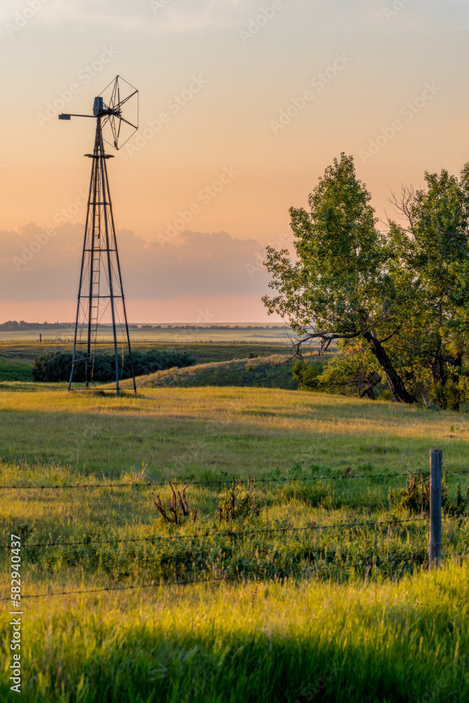 Obraz premium An old windmill in a pasture at sunset with trees and a fence line in the foreground- Orton effect