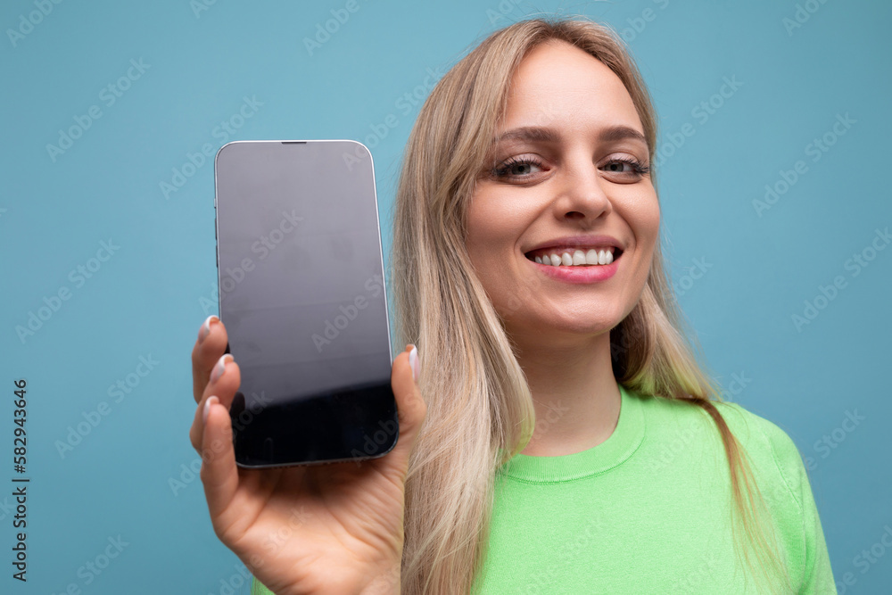 smiling bright blond girl in casual outfit holding smartphone vertically with screen forward mockup on blue background