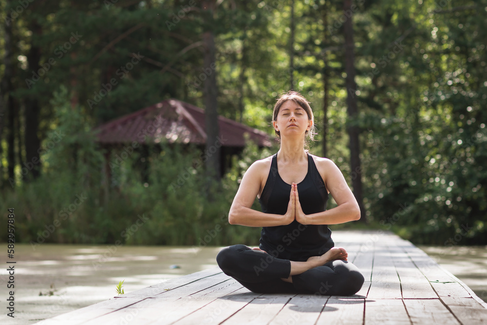 A woman leading a healthy lifestyle and practicing yoga is engaged in meditation sitting in a lotus position on a wooden path on a sunny summer morning in the park