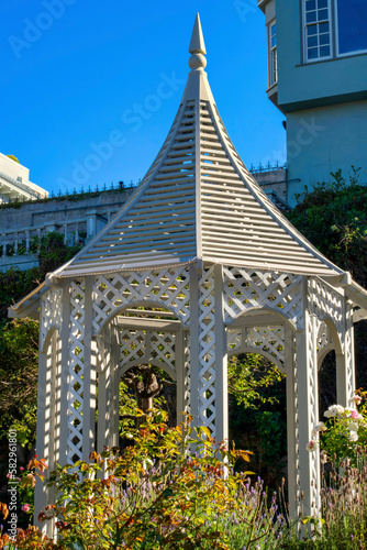 White gazebo with tall spire and top front yard trees and plants with urban or suburban background blue sky downtown