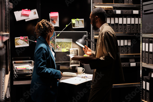 Police agents analyzing evidence and cooperating to solve crime together. African american policewoman pointing at clues hanging on detective board and policeman reading csi report photo