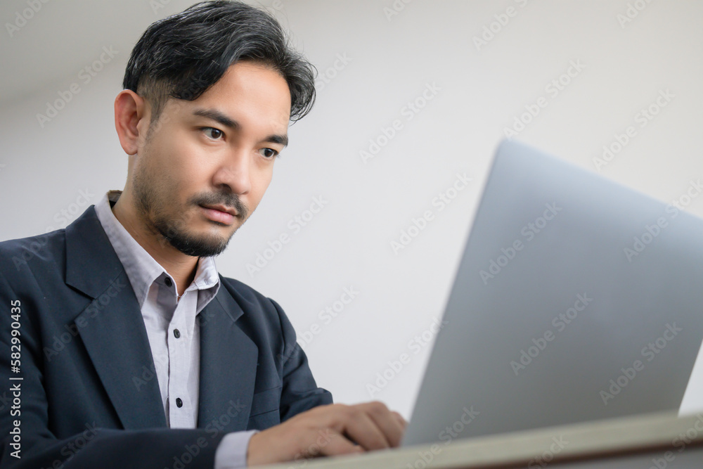 Handsome Asian business man in a suit is sitting on a laptop computer lying on a desk with a serious expression on his face. Handsome Asian business man in a suit is sitting on a laptop computer lying on a desk with a serious expression on his face.