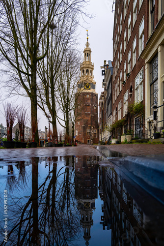Montelbaans Tower, Amsterdam downtown walkway with rain puddle reflections, traditional houses, and barren trees, on an overcast morning in the Netherlands