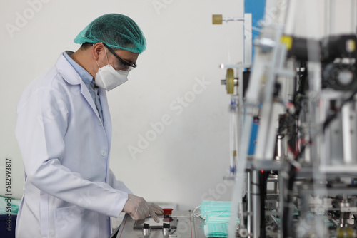 Worker in personal protective equipment or PPE inspecting quality of mask and medical face mask production line in factory  manufacturing industry and factory concept.