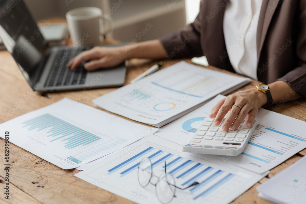 Close-up shot of businesswoman using calculator and laptop to calculate company financial result On wooden table in office and business background, tax, accounting, statistics and analytical research. Close-up shot of businesswoman using calculator and laptop to calculate company financial result On wooden table in office and business background, tax, accounting, statistics and analytical research.