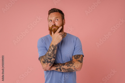 Valokuva Adult redhead bearded tattooed thoughtful man in t-shirt holding chin