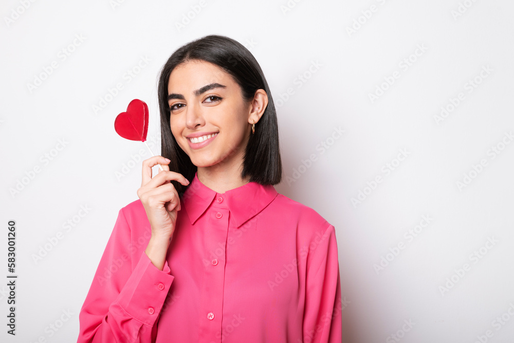 Happy young woman holding red heart shaped lollipop against white background Happy young woman holding red heart shaped lollipop against white background