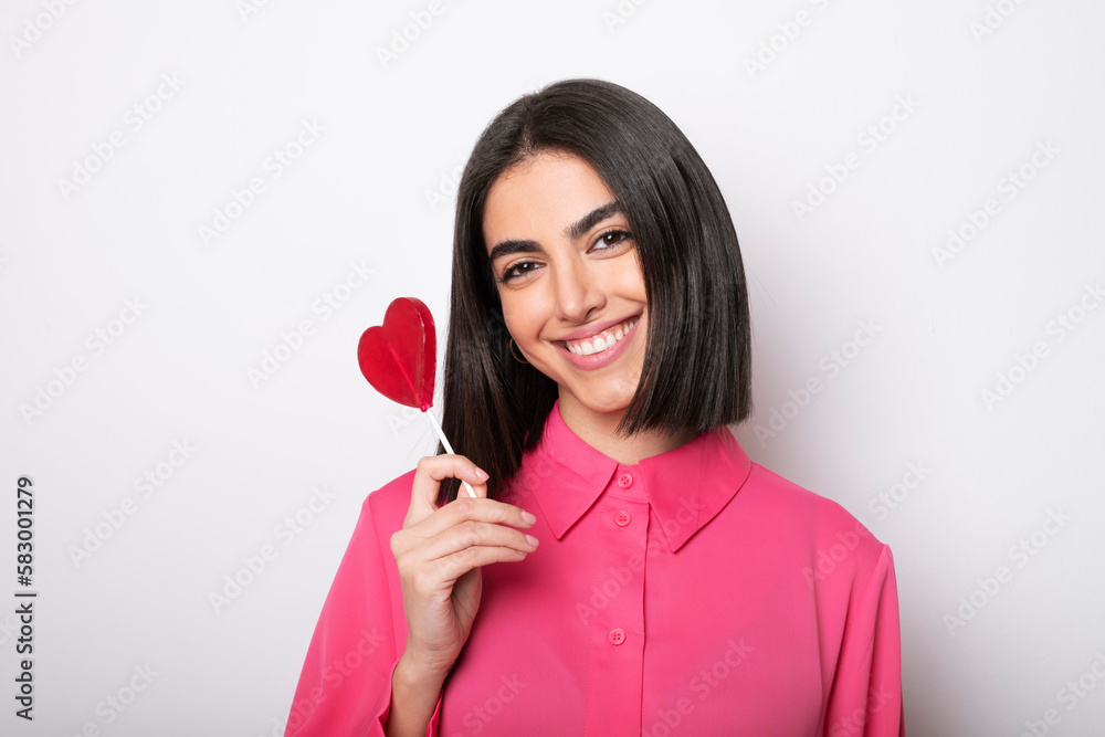 Smiling woman holding heart shaped lollipop against white background Smiling woman holding heart shaped lollipop against white background