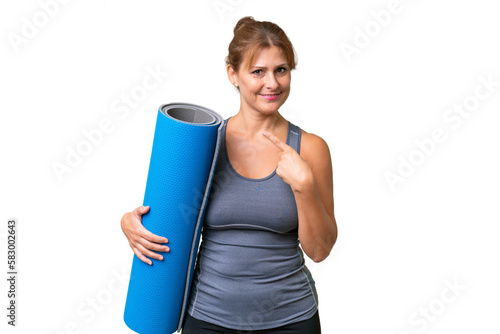 Middle-aged sport woman going to yoga classes while holding a mat over isolated background pointing to the side to present a product