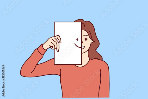 Happy woman covers half of face with paper with smiling emoticon, wanting to share good mood and positive with people around. Kind girl carelessly looks at camera enjoying life and enjoying moment 