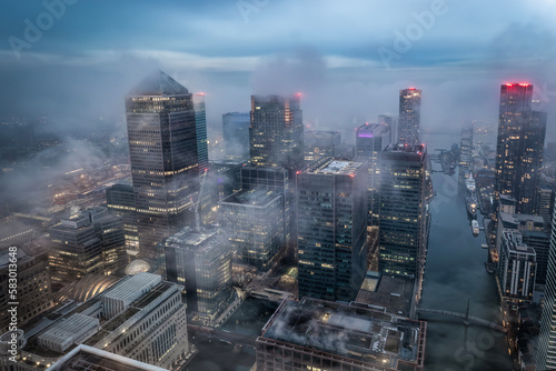 The illuminated skyline of Canary Wharf, London, during a foggy evening with the tops of the skyscrapers looking out of the clouds