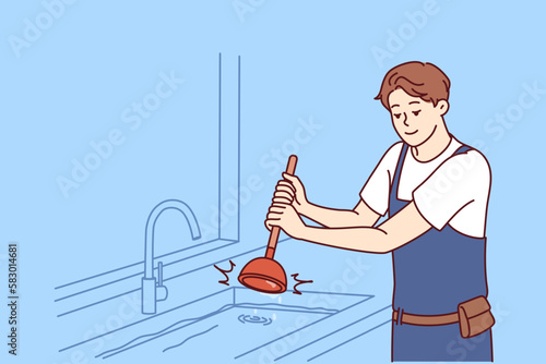 Man plumber uses plunger to clear blockage in sink in kitchen or bathroom. Guy working as plumber is dressed in overalls and is engaged in elimination breakdown associated with debris stuck in pipe 