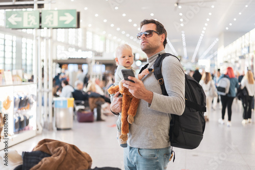 Father traveling with child, holding his infant baby boy at airport terminal, checking flight schedule, waiting to board a plane. Travel with kids concept