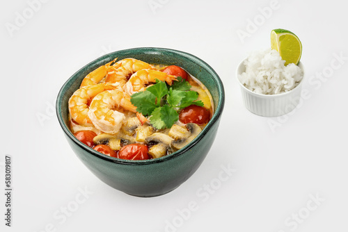 Spicy creamy soup with shrimps, cherry tomato, cilantro leaves, mushrooms in ceramic green bowl and little bowl of rice with slice of lime on light background. Asian Thai food