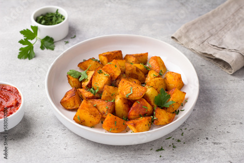 Traditional Spanish potato, patatas bravas with smoked paprika, spicy tomato sauce and parsley in a white bowl on gray stone background, top view. Delicious homemade food