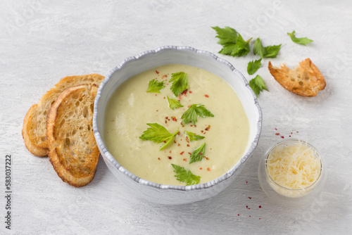 Cream soup of stalked celery with cream, garnished with celery leaves, pink pepper and served with crispy toast on a light gray background, top view