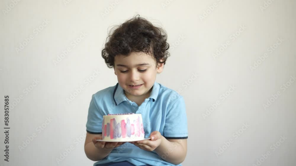 Curly cute caucasian boy holds birthday cake against white wall eats pastel coloured cake home. Childhood, holiday, sweets. Hispanic kid taste cake. Curly cute caucasian boy holds birthday cake against white wall eats pastel coloured cake home. Childhood, holiday, sweets. Hispanic kid taste cake.