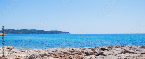 A couple standup paddleboarding on the beautiful blue sea under a clear summer sky, enjoying a tranquil day of leisure and recreation. Panoramic view.