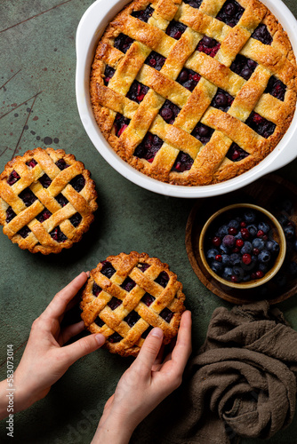 Girls hands holding homemade baked mini mixed berry crust pie or tart with lattice. Homemade, fresh berries. Green table surface, top view, vertical image.