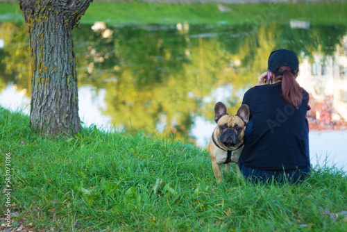 A girl is sitting on the bank of the river talking on the phone, and a French bulldog is standing next to her