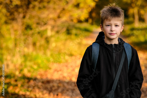 Front view of a smiling boy outdoors in an open space on a yellow, orange background with a backpack in a black jacket. caucasian blond, young guy stands with a smile, looks straight ahead, free space