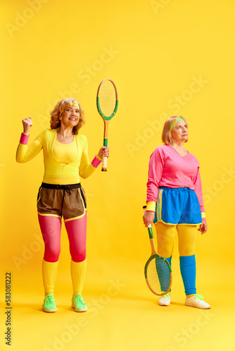 Two middle-aged, elderly women, posing in colorful uniform with tennis rackets, training against yellow studio background. Concept of sportive lifestyle, retirement, health care, wellness. Ad © master1305