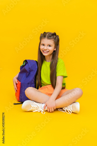 A child with a school backpack. A little girl in shorts is sitting and preparing for summer extra classes. Children's education. Yellow isolated background.