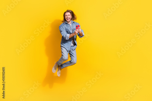 Full length photo of funny excited man wear denim jacket jumping high typing modern device isolated yellow color background
