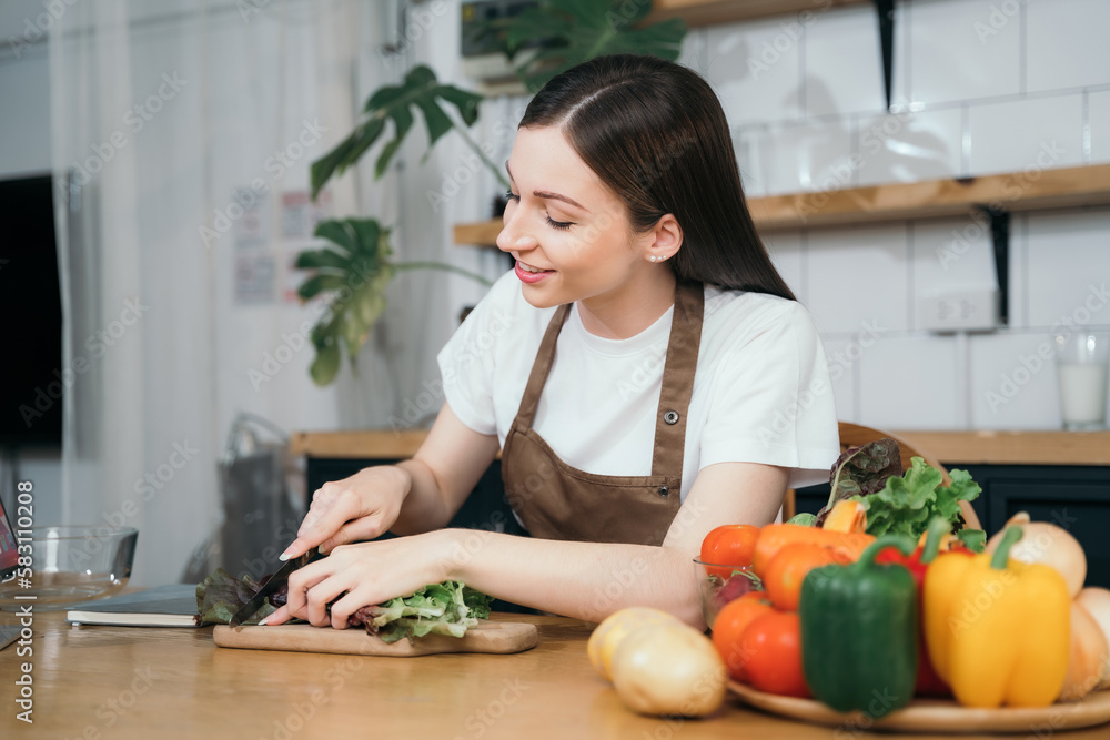 Young beautiful woman using laptop computer searching and learning for cooking healthy food from fresh vegetables and fruits in kitchen room.