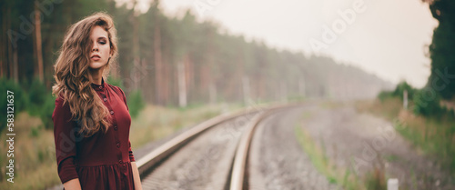 Beautiful girl with curly natural hair enjoy nature in forest on railway. Dreamer lady in burgundy dress walk on railroad. Female portrait of inspired girl on rails at dawn. Sun in hair in autumn.