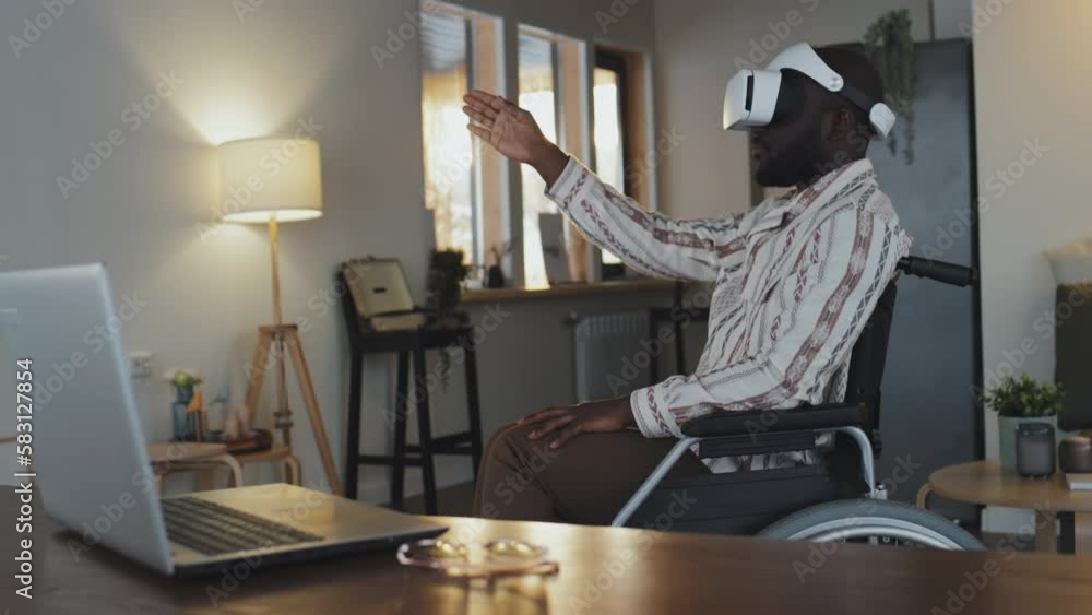 Medium full shot of black man in wheelchair using VR headset, managing some operations by means of his hand, sitting in front of his notebook in light living room at daytime Medium full shot of black man in wheelchair using VR headset, managing some operations by means of his hand, sitting in front of his notebook in light living room at daytime