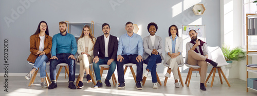Portrait of group of people sitting in row on chairs and who are listeners or candidates. Happy multiracial men and women of all ages in office sitting on chairs and looking at camera. Banner.