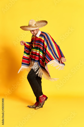 Принти на полотні Portrait of young handsome man in colorful festive clothes, poncho and sombrero posing, dancing against yellow studio background