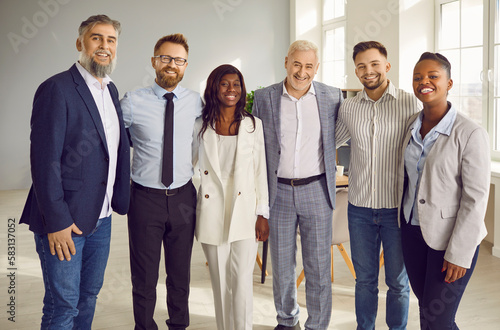 Group of business multiethnic people men and women looking at camera hugging and smiling indoors. Company employees team or group of staff standing in the office confidently and cheerfully.