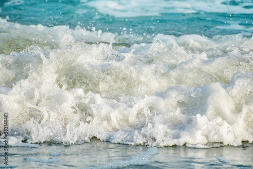 Fototapeta Naklejka Na Ścianę i Meble -  Close-up of the sea wave with the foam surging on the shore. View of sea water in soft focus. Background in a light blur on the theme of a serene summer vacation and relaxed vacation.