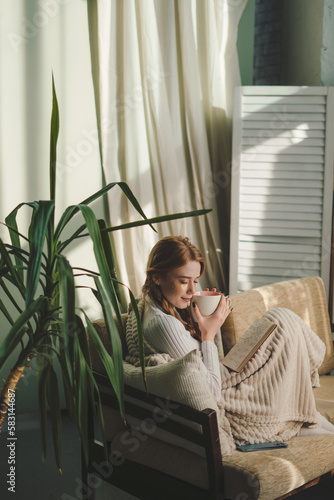 Indoor shot of young woman sitting on sofa covered with a blanket in the modern living room, holding cup. Notepads and documents. Creative artwork. © Strelciuc