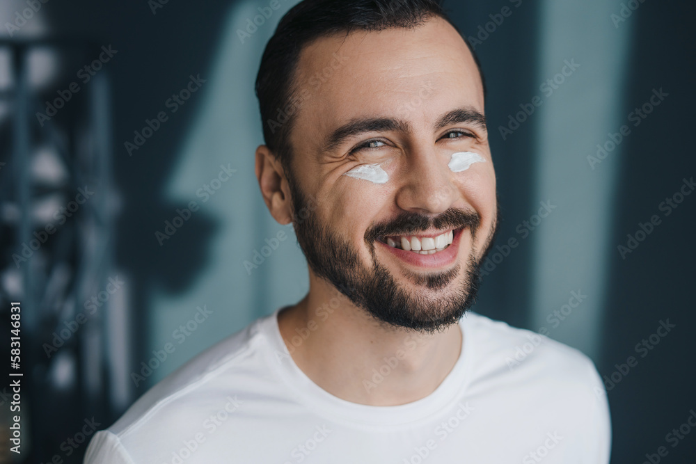 Closeup portrait of handsome bearded man with moisturising cream on cheeks, looking and smiling at camera. Beauty portrait. Facial treatment. Closeup portrait Closeup portrait of handsome bearded man with moisturising cream on cheeks, looking and smiling at camera. Beauty portrait. Facial treatment. Closeup portrait