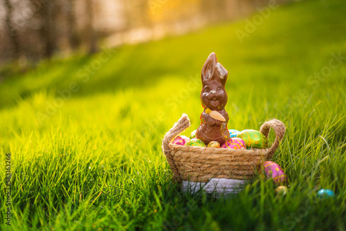 Easter eggs in basket with easter bunny on top. Chocolate rabbit with colorful decorated eggs in wicker basket in grass. Magical morning light, spring season holidays. Traditional egg hunt