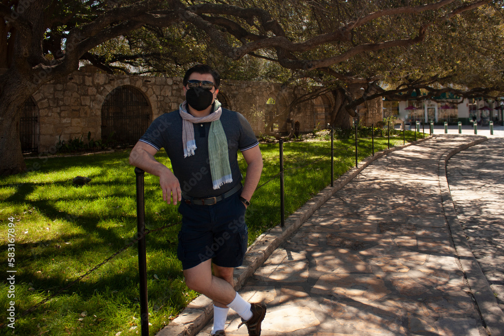 Fototapeta premium Caucasian Mexican man visitor wearing mask and sunglasses on Alamo sightseeing in San Antonio Texas on street next to the garden